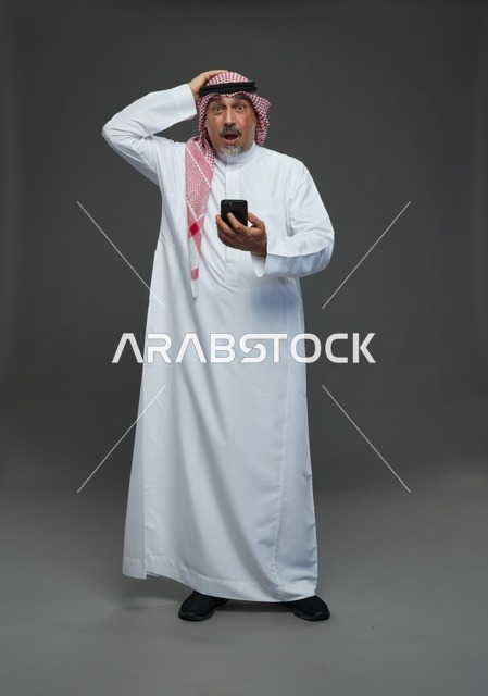 رجل  عربي سعودي خليجي مسن يرتدي الزي الثوب الابيض والشماغ ويمسك الهاتف المحمول,  ايماءات واشارات مختلفه , كامل الجسم, خلفيه رماديه