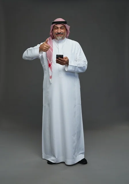 رجل  عربي سعودي خليجي مسن يرتدي الزي الثوب الابيض والشماغ ويمسك الهاتف المحمول,  ايماءات واشارات مختلفه , كامل الجسم, خلفيه رماديه