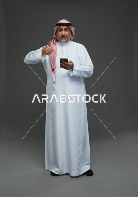 رجل  عربي سعودي خليجي مسن يرتدي الزي الثوب الابيض والشماغ ويمسك الهاتف المحمول,  ايماءات واشارات مختلفه , كامل الجسم, خلفيه رماديه