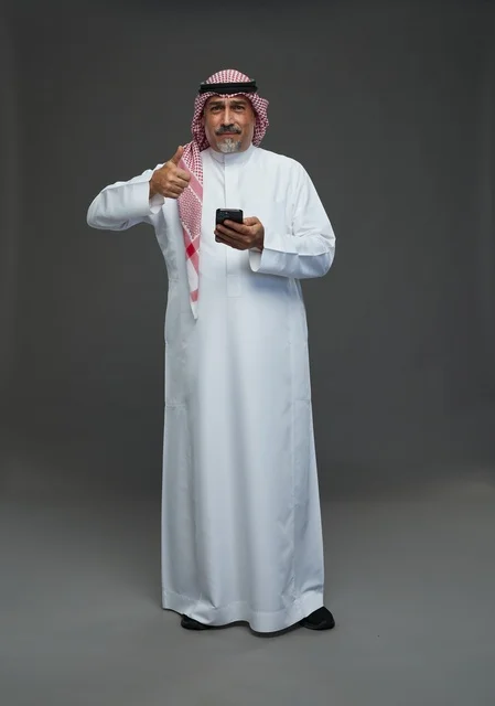 رجل  عربي سعودي خليجي مسن يرتدي الزي الثوب الابيض والشماغ ويمسك الهاتف المحمول,  ايماءات واشارات مختلفه , كامل الجسم, خلفيه رماديه