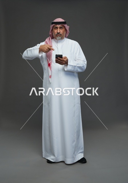 رجل  عربي سعودي خليجي مسن يرتدي الزي الثوب الابيض والشماغ ويمسك الهاتف المحمول,  ايماءات واشارات مختلفه , كامل الجسم, خلفيه رماديه