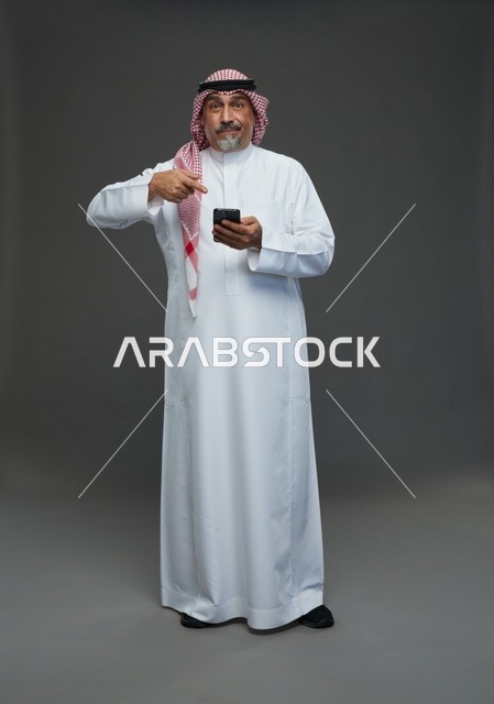 رجل  عربي سعودي خليجي مسن يرتدي الزي الثوب الابيض والشماغ ويمسك الهاتف المحمول,  ايماءات واشارات مختلفه , كامل الجسم, خلفيه رماديه