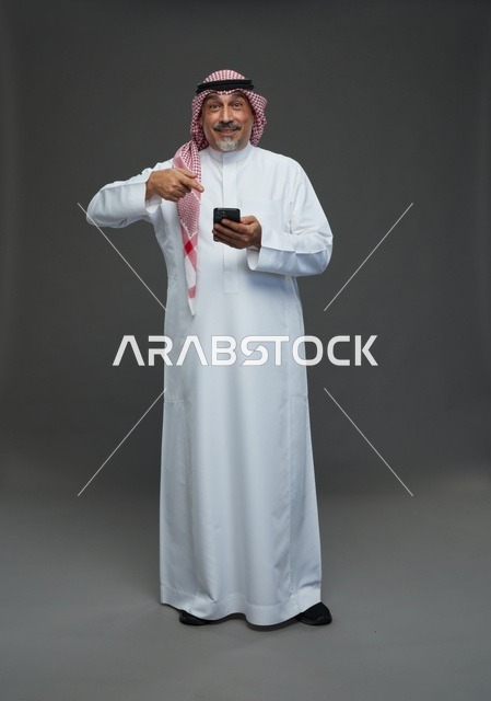 رجل  عربي سعودي خليجي مسن يرتدي الزي الثوب الابيض والشماغ ويمسك الهاتف المحمول,  ايماءات واشارات مختلفه , كامل الجسم, خلفيه رماديه