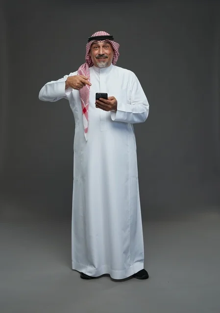 رجل  عربي سعودي خليجي مسن يرتدي الزي الثوب الابيض والشماغ ويمسك الهاتف المحمول,  ايماءات واشارات مختلفه , كامل الجسم, خلفيه رماديه