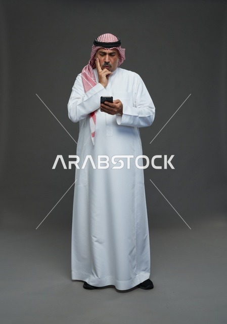 رجل  عربي سعودي خليجي مسن يرتدي الزي الثوب الابيض والشماغ ويمسك الهاتف المحمول,  ايماءات واشارات مختلفه , كامل الجسم, خلفيه رماديه