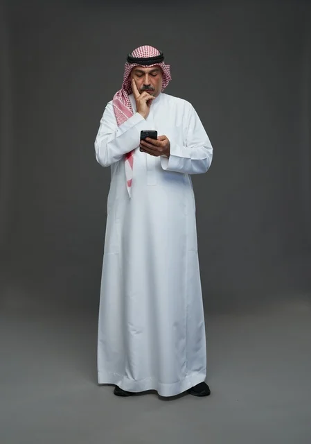 An elderly Arab Saudi Gulf man wearing a white thobe and a shemagh, holding a mobile phone, with various gestures and signals, full body, gray background.