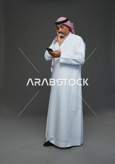 رجل  عربي سعودي خليجي مسن يرتدي الزي الثوب الابيض والشماغ ويمسك الهاتف المحمول,  ايماءات واشارات مختلفه , كامل الجسم, خلفيه رماديه