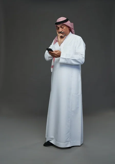 رجل  عربي سعودي خليجي مسن يرتدي الزي الثوب الابيض والشماغ ويمسك الهاتف المحمول,  ايماءات واشارات مختلفه , كامل الجسم, خلفيه رماديه