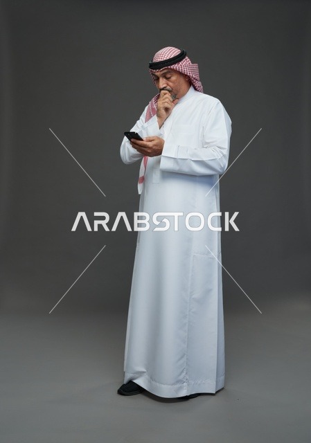 رجل  عربي سعودي خليجي مسن يرتدي الزي الثوب الابيض والشماغ ويمسك الهاتف المحمول,  ايماءات واشارات مختلفه , كامل الجسم, خلفيه رماديه