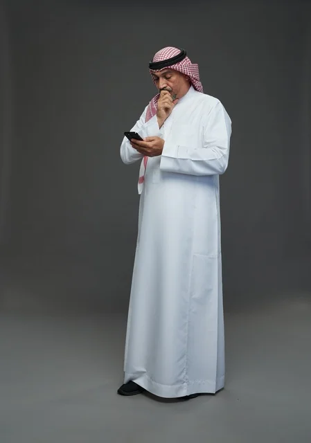 An elderly Arab Saudi Gulf man wearing a white thobe and a shemagh, holding a mobile phone, with various gestures and signals, full body, gray background.