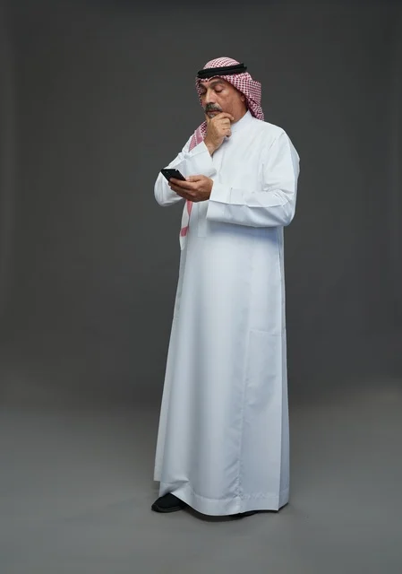 رجل  عربي سعودي خليجي مسن يرتدي الزي الثوب الابيض والشماغ ويمسك الهاتف المحمول,  ايماءات واشارات مختلفه , كامل الجسم, خلفيه رماديه