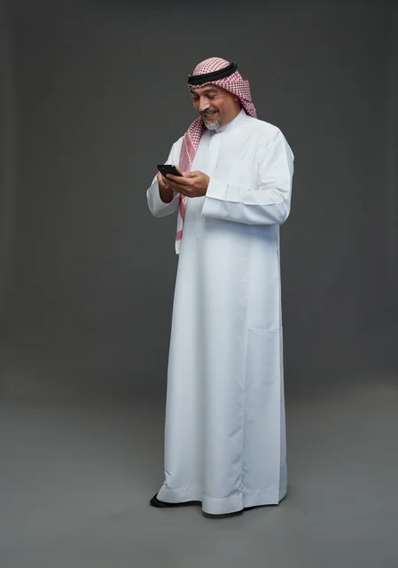رجل  عربي سعودي خليجي مسن يرتدي الزي الثوب الابيض والشماغ ويمسك الهاتف المحمول,  ايماءات واشارات مختلفه , كامل الجسم, خلفيه رماديه