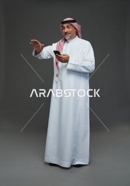 رجل  عربي سعودي خليجي مسن يرتدي الزي الثوب الابيض والشماغ ويمسك الهاتف المحمول,  ايماءات واشارات مختلفه , كامل الجسم, خلفيه رماديه