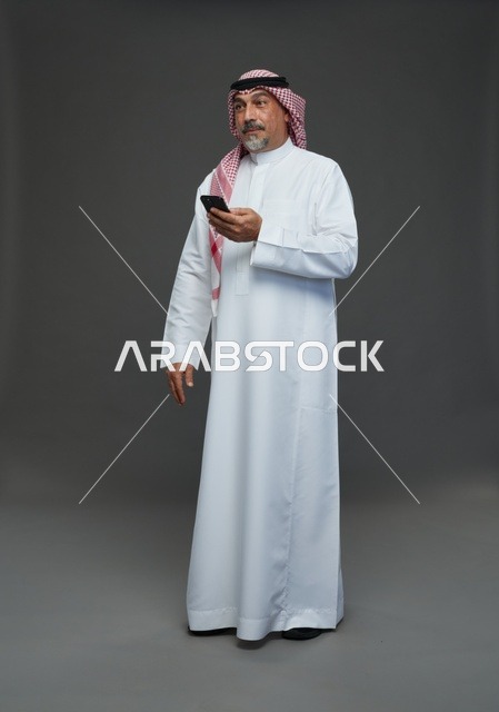 رجل  عربي سعودي خليجي مسن يرتدي الزي الثوب الابيض والشماغ ويمسك الهاتف المحمول,  ايماءات واشارات مختلفه , كامل الجسم, خلفيه رماديه