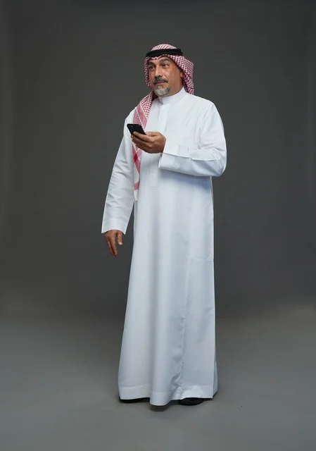 An elderly Arab Saudi Gulf man wearing a white thobe and a shemagh, holding a mobile phone, with various gestures and signals, full body, gray background.