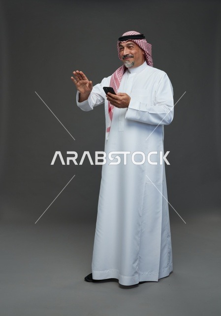 رجل  عربي سعودي خليجي مسن يرتدي الزي الثوب الابيض والشماغ ويمسك الهاتف المحمول,  ايماءات واشارات مختلفه , كامل الجسم, خلفيه رماديه