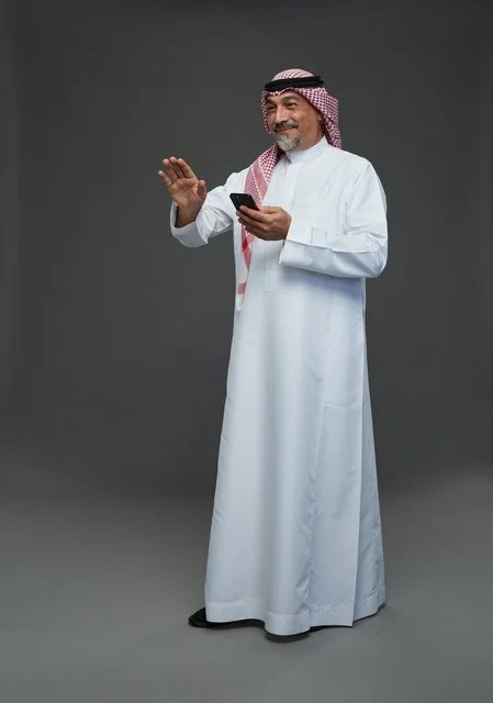 رجل  عربي سعودي خليجي مسن يرتدي الزي الثوب الابيض والشماغ ويمسك الهاتف المحمول,  ايماءات واشارات مختلفه , كامل الجسم, خلفيه رماديه