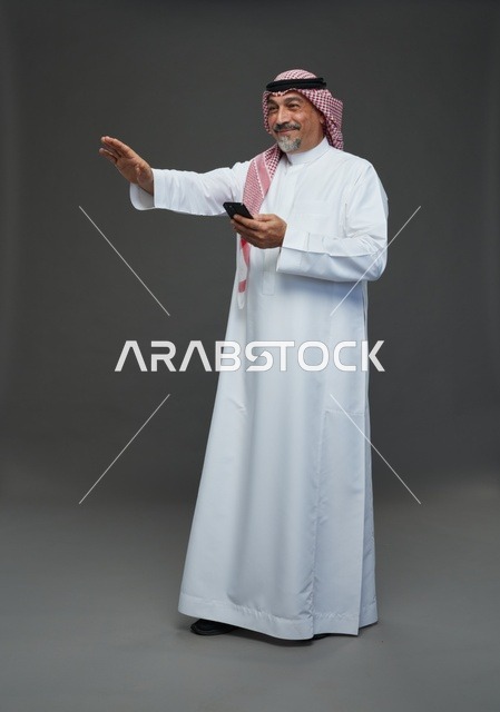 رجل  عربي سعودي خليجي مسن يرتدي الزي الثوب الابيض والشماغ ويمسك الهاتف المحمول,  ايماءات واشارات مختلفه , كامل الجسم, خلفيه رماديه