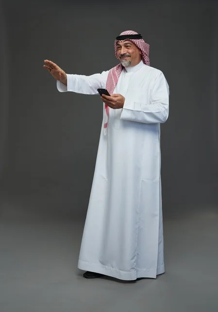 رجل  عربي سعودي خليجي مسن يرتدي الزي الثوب الابيض والشماغ ويمسك الهاتف المحمول,  ايماءات واشارات مختلفه , كامل الجسم, خلفيه رماديه