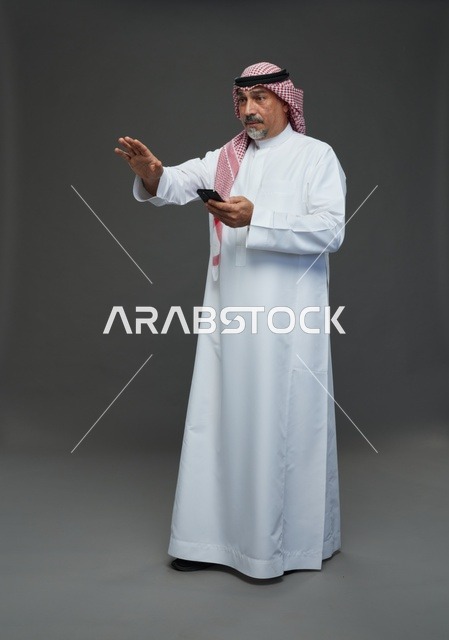 رجل  عربي سعودي خليجي مسن يرتدي الزي الثوب الابيض والشماغ ويمسك الهاتف المحمول,  ايماءات واشارات مختلفه , كامل الجسم, خلفيه رماديه