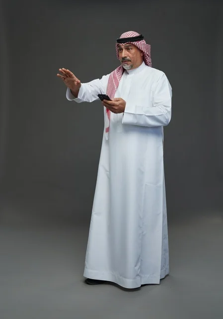 An elderly Arab Saudi Gulf man wearing a white thobe and a shemagh, holding a mobile phone, with various gestures and signals, full body, gray background.