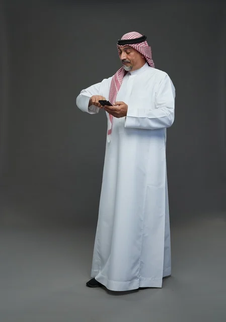 An elderly Arab Saudi Gulf man wearing a white thobe and a shemagh, holding a mobile phone, with various gestures and signals, full body, gray background.