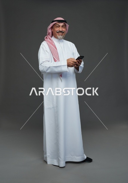 رجل  عربي سعودي خليجي مسن يرتدي الزي الثوب الابيض والشماغ ويمسك الهاتف المحمول,  ايماءات واشارات مختلفه , كامل الجسم, خلفيه رماديه