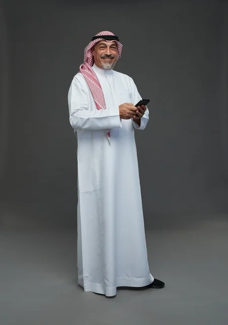 رجل  عربي سعودي خليجي مسن يرتدي الزي الثوب الابيض والشماغ ويمسك الهاتف المحمول,  ايماءات واشارات مختلفه , كامل الجسم, خلفيه رماديه