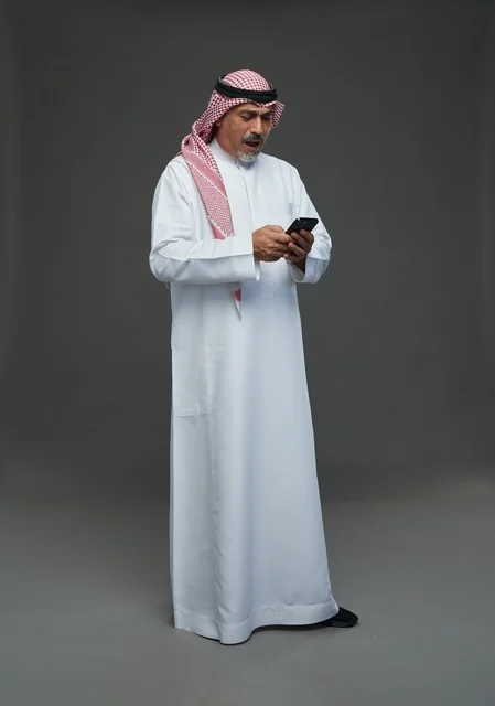 An elderly Arab Saudi Gulf man wearing a white thobe and a shemagh, holding a mobile phone, with various gestures and signals, full body, gray background.