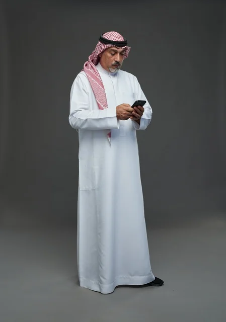An elderly Arab Saudi Gulf man wearing a white thobe and a shemagh, holding a mobile phone, with various gestures and signals, full body, gray background.