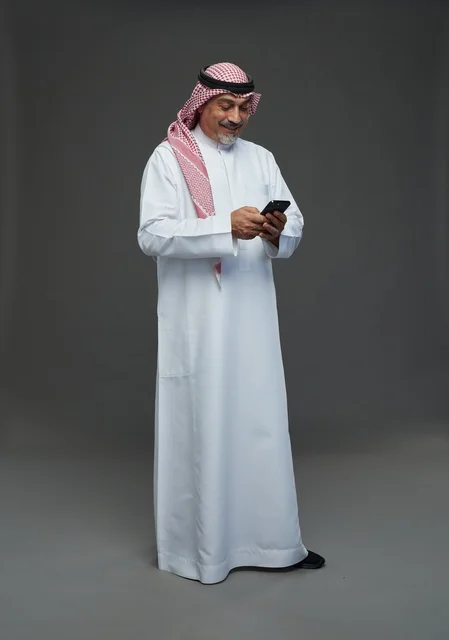 An elderly Arab Saudi Gulf man wearing a white thobe and a shemagh, holding a mobile phone, with various gestures and signals, full body, gray background.