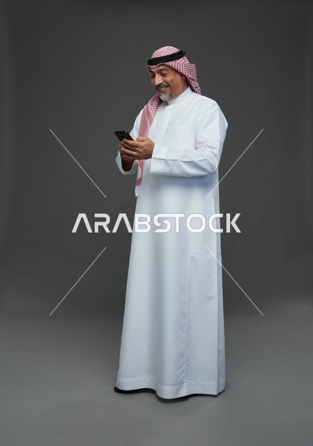 رجل  عربي سعودي خليجي مسن يرتدي الزي الثوب الابيض والشماغ ويمسك الهاتف المحمول,  ايماءات واشارات مختلفه , كامل الجسم, خلفيه رماديه