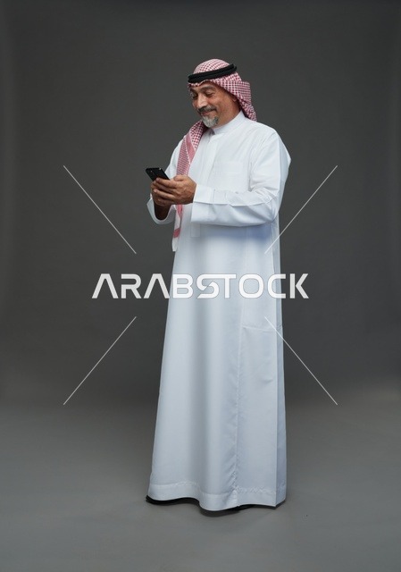 رجل  عربي سعودي خليجي مسن يرتدي الزي الثوب الابيض والشماغ ويمسك الهاتف المحمول,  ايماءات واشارات مختلفه , كامل الجسم, خلفيه رماديه