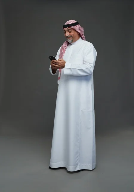 رجل  عربي سعودي خليجي مسن يرتدي الزي الثوب الابيض والشماغ ويمسك الهاتف المحمول,  ايماءات واشارات مختلفه , كامل الجسم, خلفيه رماديه