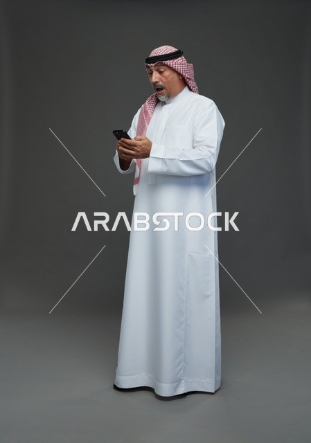 رجل  عربي سعودي خليجي مسن يرتدي الزي الثوب الابيض والشماغ ويمسك الهاتف المحمول,  ايماءات واشارات مختلفه , كامل الجسم, خلفيه رماديه