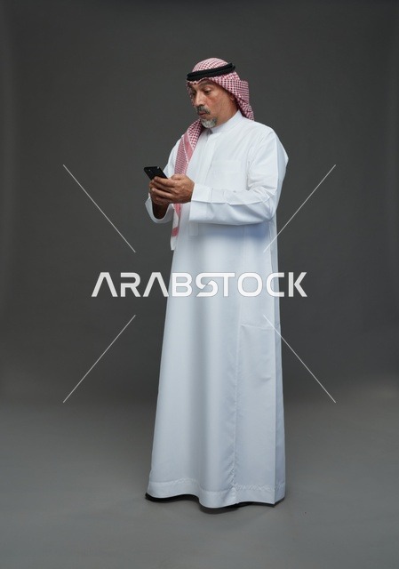 رجل  عربي سعودي خليجي مسن يرتدي الزي الثوب الابيض والشماغ ويمسك الهاتف المحمول,  ايماءات واشارات مختلفه , كامل الجسم, خلفيه رماديه
