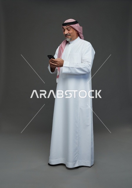 رجل  عربي سعودي خليجي مسن يرتدي الزي الثوب الابيض والشماغ ويمسك الهاتف المحمول,  ايماءات واشارات مختلفه , كامل الجسم, خلفيه رماديه