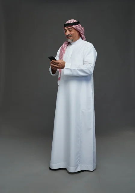 رجل  عربي سعودي خليجي مسن يرتدي الزي الثوب الابيض والشماغ ويمسك الهاتف المحمول,  ايماءات واشارات مختلفه , كامل الجسم, خلفيه رماديه