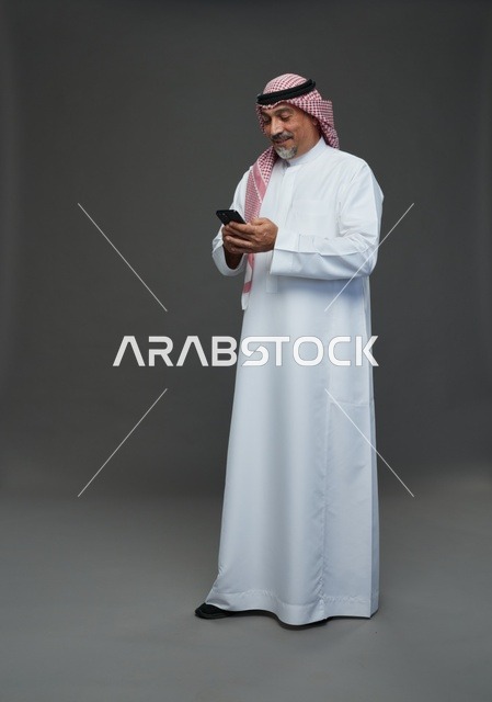 رجل  عربي سعودي خليجي مسن يرتدي الزي الثوب الابيض والشماغ ويمسك الهاتف المحمول,  ايماءات واشارات مختلفه , كامل الجسم, خلفيه رماديه
