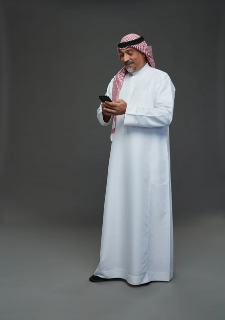 An elderly Arab Saudi Gulf man wearing a white thobe and a shemagh, holding a mobile phone, with various gestures and signals, full body, gray background.