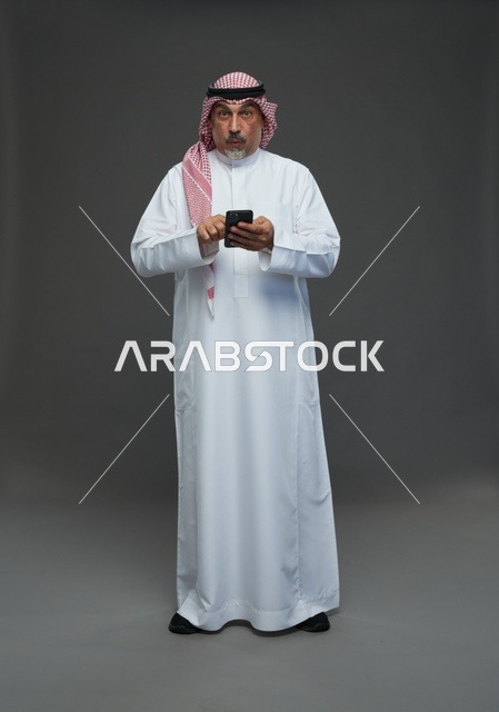 رجل  عربي سعودي خليجي مسن يرتدي الزي الثوب الابيض والشماغ ويمسك الهاتف المحمول,  ايماءات واشارات مختلفه , كامل الجسم, خلفيه رماديه