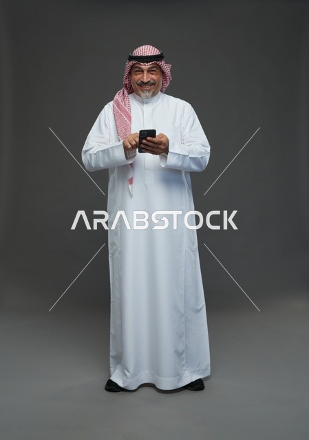 رجل  عربي سعودي خليجي مسن يرتدي الزي الثوب الابيض والشماغ ويمسك الهاتف المحمول,  ايماءات واشارات مختلفه , كامل الجسم, خلفيه رماديه