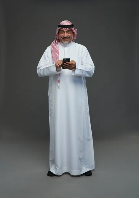 رجل  عربي سعودي خليجي مسن يرتدي الزي الثوب الابيض والشماغ ويمسك الهاتف المحمول,  ايماءات واشارات مختلفه , كامل الجسم, خلفيه رماديه
