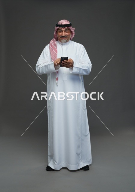 رجل  عربي سعودي خليجي مسن يرتدي الزي الثوب الابيض والشماغ ويمسك الهاتف المحمول,  ايماءات واشارات مختلفه , كامل الجسم, خلفيه رماديه