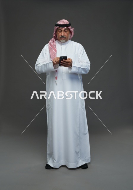 رجل  عربي سعودي خليجي مسن يرتدي الزي الثوب الابيض والشماغ ويمسك الهاتف المحمول,  ايماءات واشارات مختلفه , كامل الجسم, خلفيه رماديه