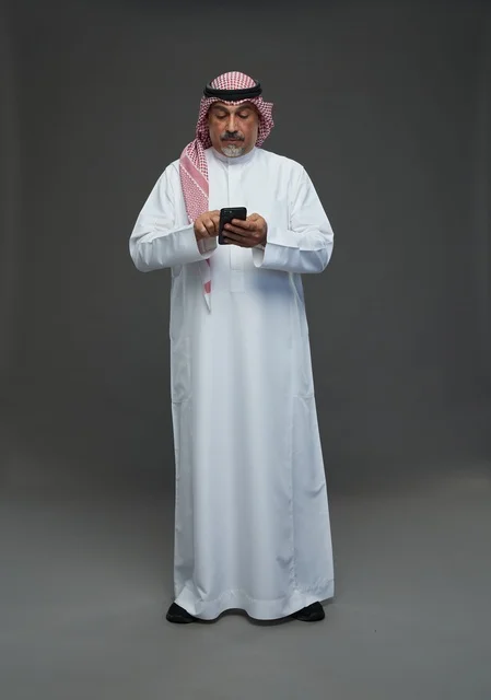 رجل  عربي سعودي خليجي مسن يرتدي الزي الثوب الابيض والشماغ ويمسك الهاتف المحمول,  ايماءات واشارات مختلفه , كامل الجسم, خلفيه رماديه