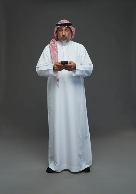 An elderly Arab Saudi Gulf man wearing a white thobe and a shemagh, holding a mobile phone, with various gestures and signals, full body, gray background.