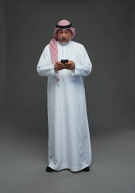 رجل  عربي سعودي خليجي مسن يرتدي الزي الثوب الابيض والشماغ ويمسك الهاتف المحمول,  ايماءات واشارات مختلفه , كامل الجسم, خلفيه رماديه