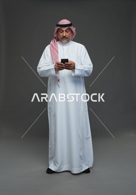 رجل  عربي سعودي خليجي مسن يرتدي الزي الثوب الابيض والشماغ ويمسك الهاتف المحمول,  ايماءات واشارات مختلفه , كامل الجسم, خلفيه رماديه