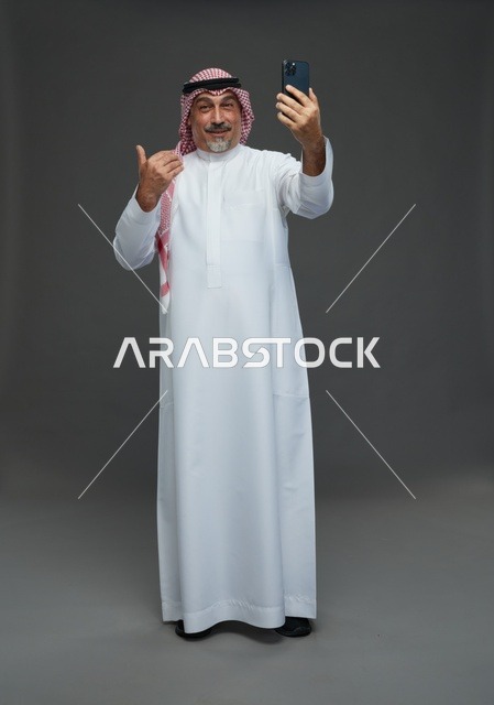 رجل  عربي سعودي خليجي مسن يرتدي الزي الثوب الابيض والشماغ ويمسك الهاتف المحمول, التقاط صورة سيلفي, اجراء مكالمة فيديو عبر الجوال,  ايماءات واشارات مختلفه , كامل الجسم, خلفيه رماديه